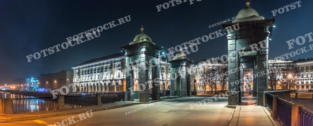 На фотографии изображён мост Ломоносова в Санкт-Петербурге, снятый ночью. Мост, освещённый уличными фонарями, пересекает реку Фонтанку и отличается своими выразительными башенками с куполами. Ночные огни добавляют магическую атмосферу к сцене, а река Фонтанка тянется вдоль, отражая свет. Это изображение подойдёт для статей о городской архитектуре и истории, а также может быть использовано в туристических проспектах и фотоальбомах.